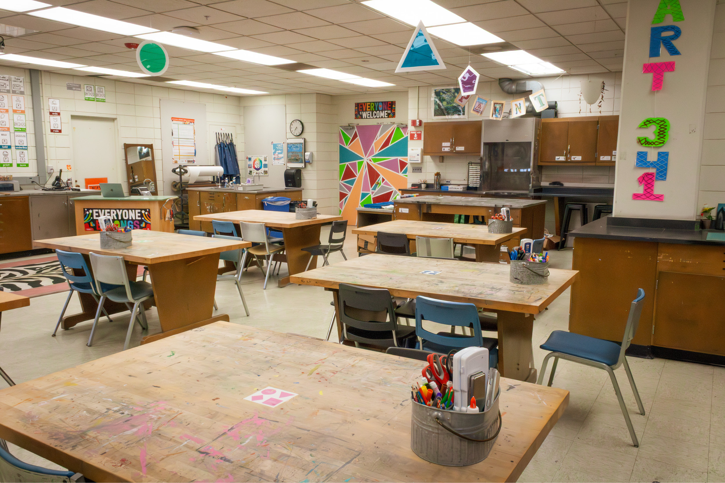 Overview of the art education classroom. There are multiple workstations, each with a caddy filled with art supplies. Fun posters are displayed on the walls.