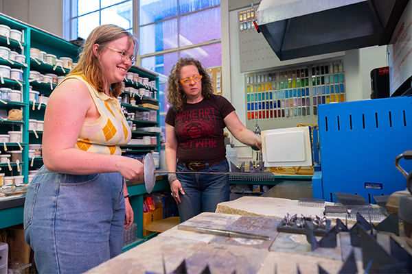 Two metalsmithing students smile as they insert their project into a kiln.