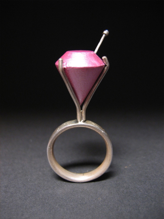 A handcrafted metallic ring inset with a tall, fake, pink diamond with a pin sticking out of it. 