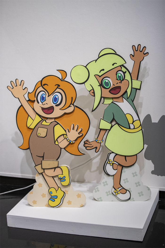 Cardboard cutouts of two cartoon girls. The girl on the left has red hair and is wearing a yellow shirt, sneakers, and brown overalls. The girl on the right has mint green hair and is wearing a green floral outfit. 