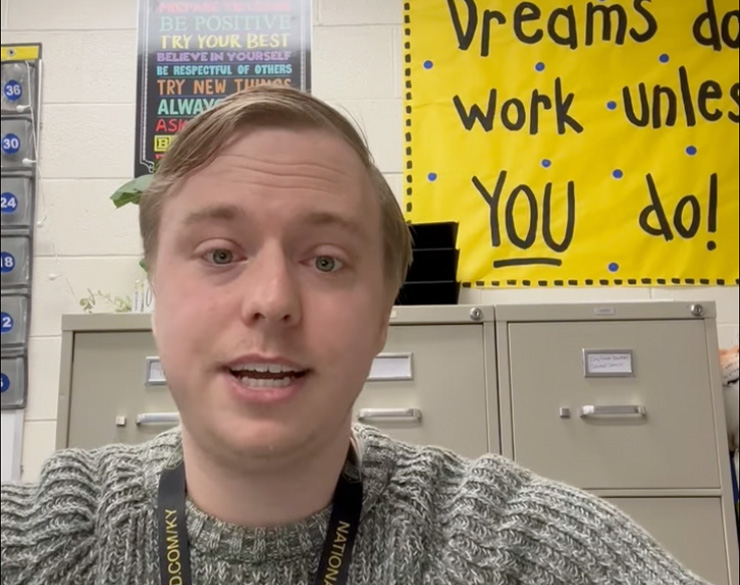 A blond man in a gray sweater, sitting in front of filing cabinets under inspirational posters, talks to the camera.