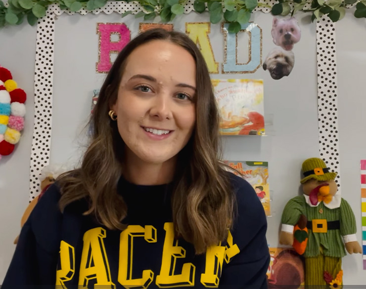 An alumnus wearing a Racer t-shirt, sitting next to a turkey plush in front of a board that says, “READ,” with books under it, smiles into the camera. 