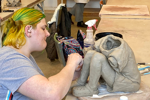 A ceramics student carves a sculpture of a seated child from clay. 