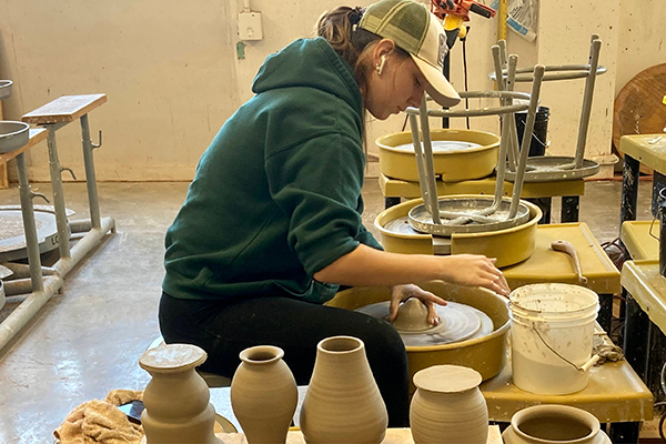 A ceramics student works at a pottery wheel; a series of ceramic vases sit on a table next to her.