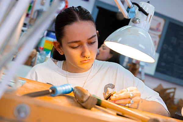 A metalsmithing student refines her wares under a lamp. 