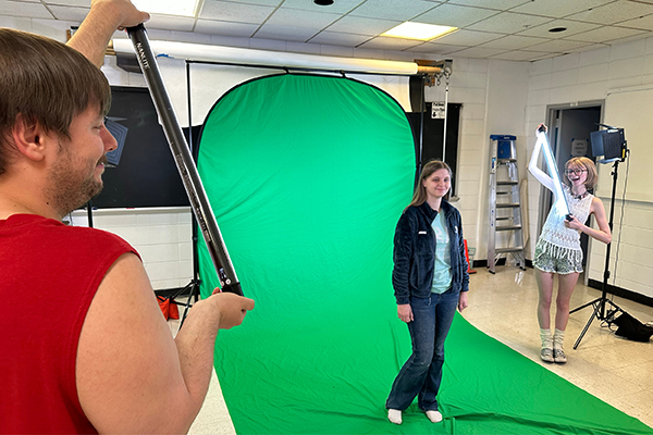 Photography students learn editorial photography by holding light bars and taking pictures of each other in front of a green screen.