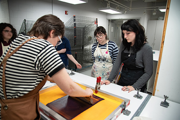 Printmaking students gather around a visiting printmaker as she inks a silkscreen. 