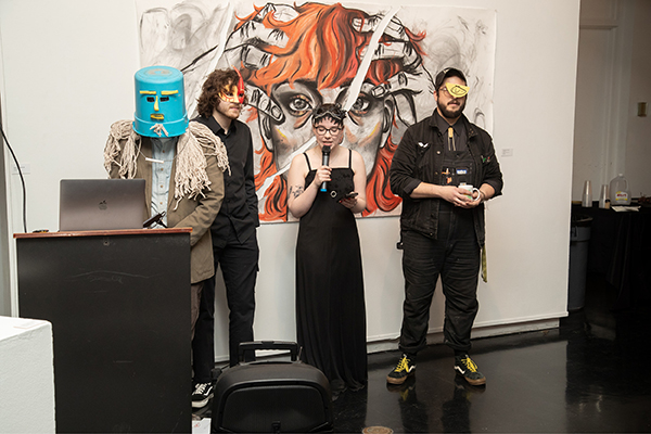 Four students give a presentation in front of a charcoal-and-pastel drawing of a stressed woman clutching her head. The woman is gray charcoal, while her hair is a bright orange pastel. Three of the students are wearing black and funky masks. The fourth student is wearing a blue bucket with a silly-face cutout over a tan jacket with mop fringe. 
