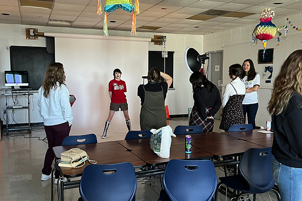 Photography students gather together as a student in green overalls takes editorial photos of another student wearing a red t-shirt, short cargo shorts, and knee-high black Converse sneakers.