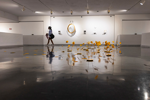 A student walks by an exhibition in the Eagle Upper Gallery, featuring a mirror on the wall and plant-like objects on the ground.