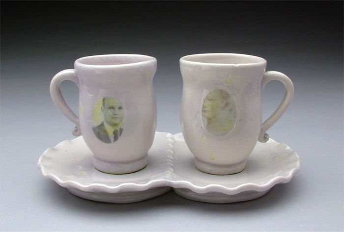 A handcrafted set of ceramic mugs sits on a plate for two. The mug on the left is adorned with the faded photo of a man, while the mug on the right has the faded photo of a woman. 