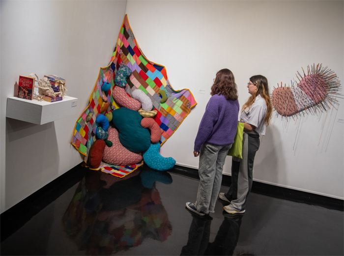 Two students look at a fabric installation of a vibrant patchwork quilt hanging from the corner, with multicolored, bean-shaped pillows spilling outward. 