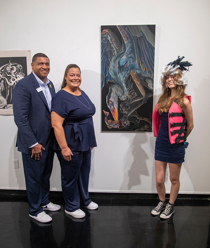 Dr. and Mrs. Patterson and a student stand next to the student’s painting of a bird catching a fish. The student wears an eclectic outfit featuring a feather headpiece, a hot pink life jacket, a navy cocktail dress, and sneakers. 
