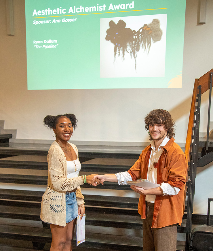 Two students holding certificates—a Black female and a white male—shake hands. Behind them, a projection reads, “Aesthetic Alchemist Award. Sponsor: Ann Gosser. Ryan Dollum. ‘The Pipeline’.”