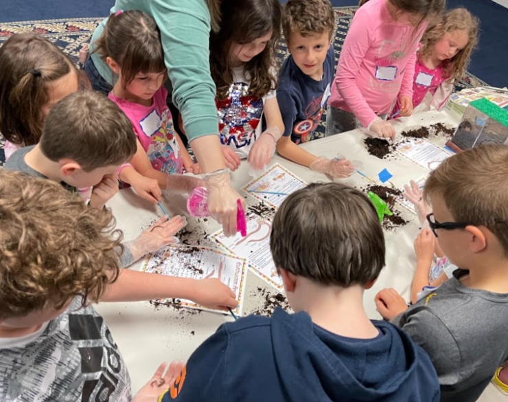 Campers stand around a table with worms and dirt while an adult sprays water mist on the worms