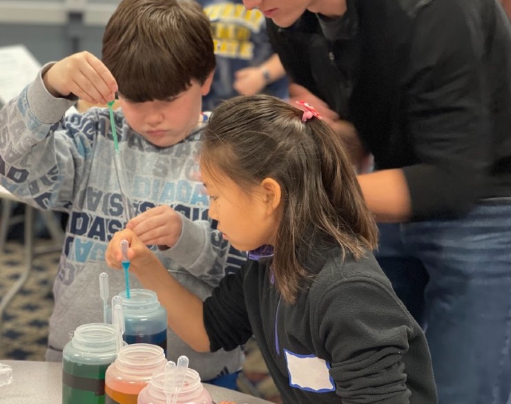 Two campers using droppers to add colored water to a container while an adult instructor helps them