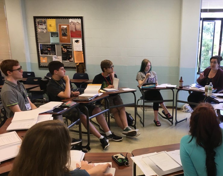 PAWW campers sitting with notebooks and pencils at desks arranged in a circle, attentatively listening to their instructor