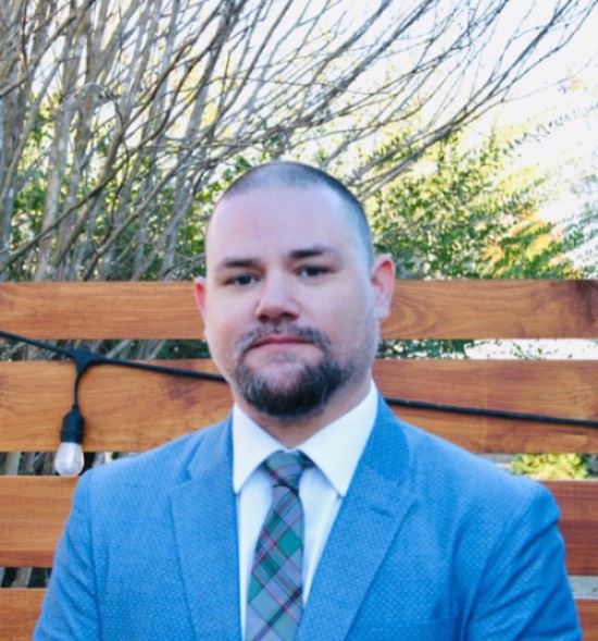 Dr. Christopher Craig in a blue jacket, white shirt and plaid tie, sitting on a wooden bench