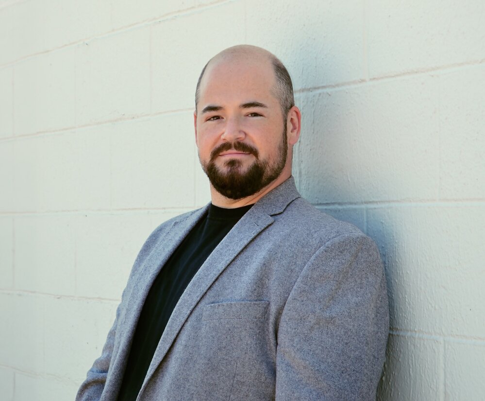 Justin Ford wearing a grey blazer leaning on wall
