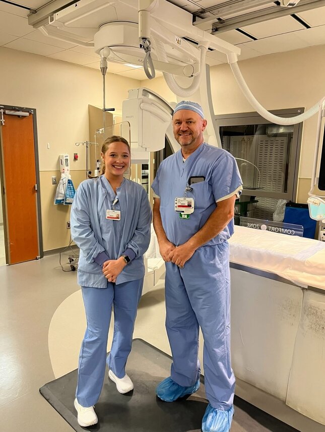 A female student nurse on the left stands next to a male RN on the right in a medical setting.