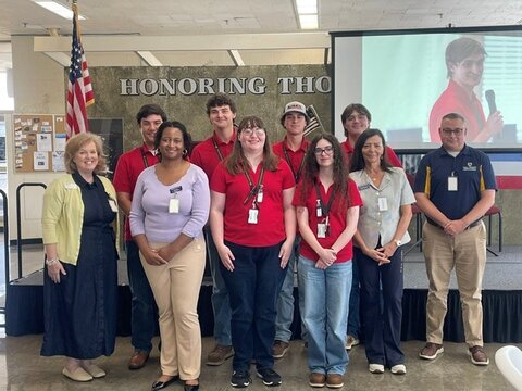A group of students in red shirts and faculty in front of a screen and an American flag.