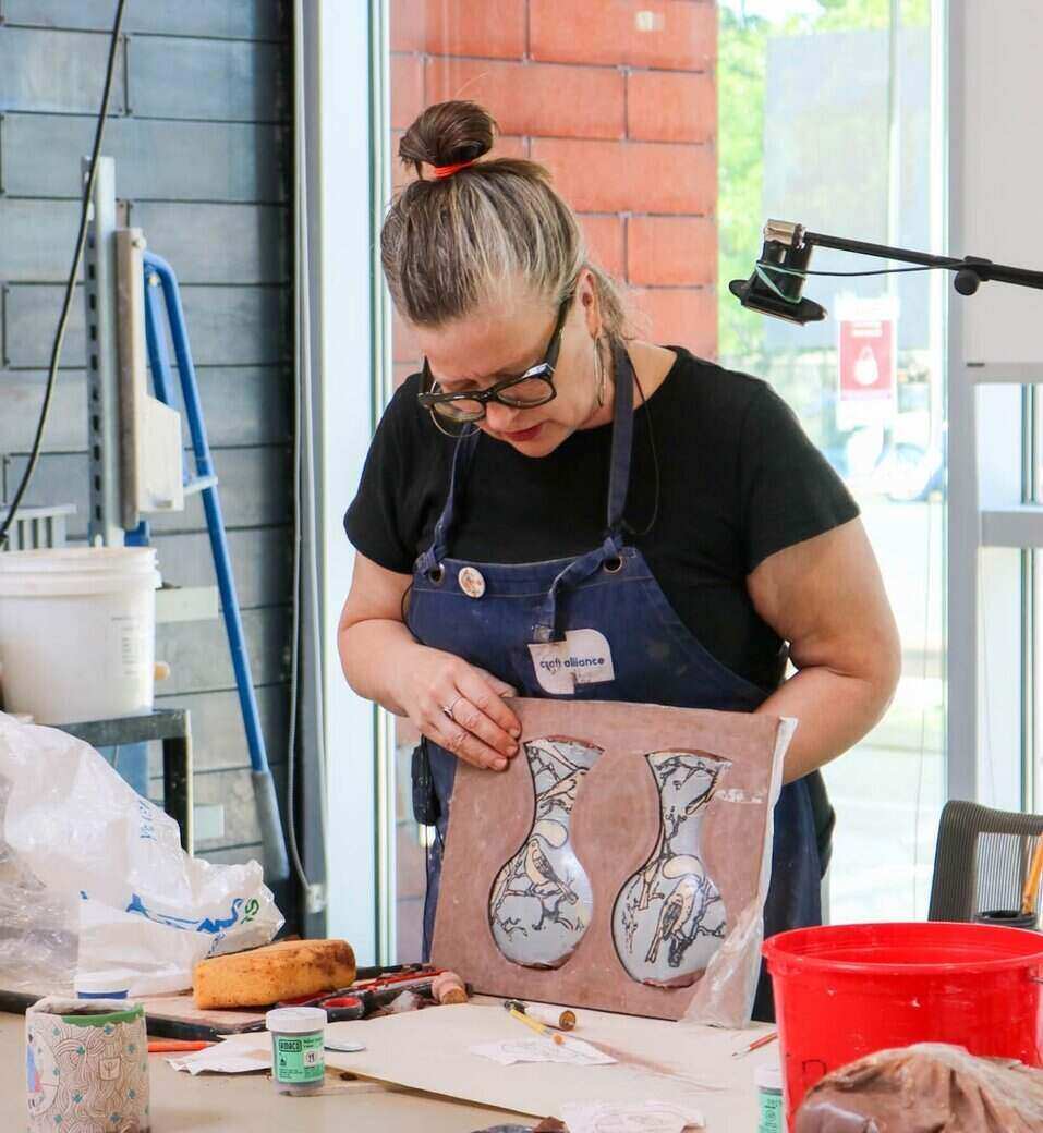Pattie Chalmers in a black shirt, denim apron and glasses displaying two ceramic vases.